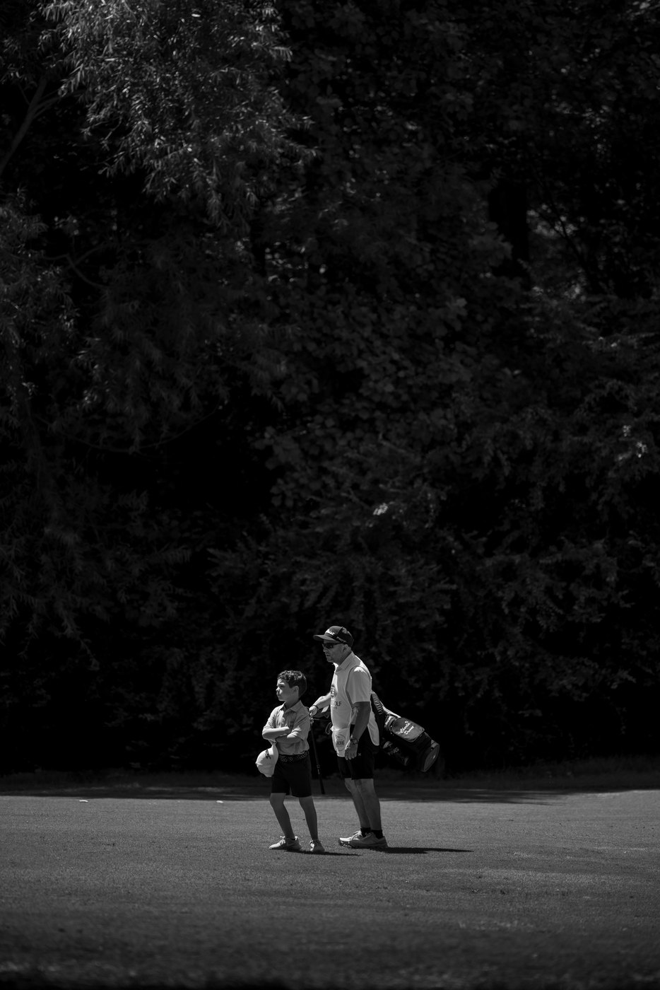 Player and caddie crossing a cart path against a wall of dark trees, black and white