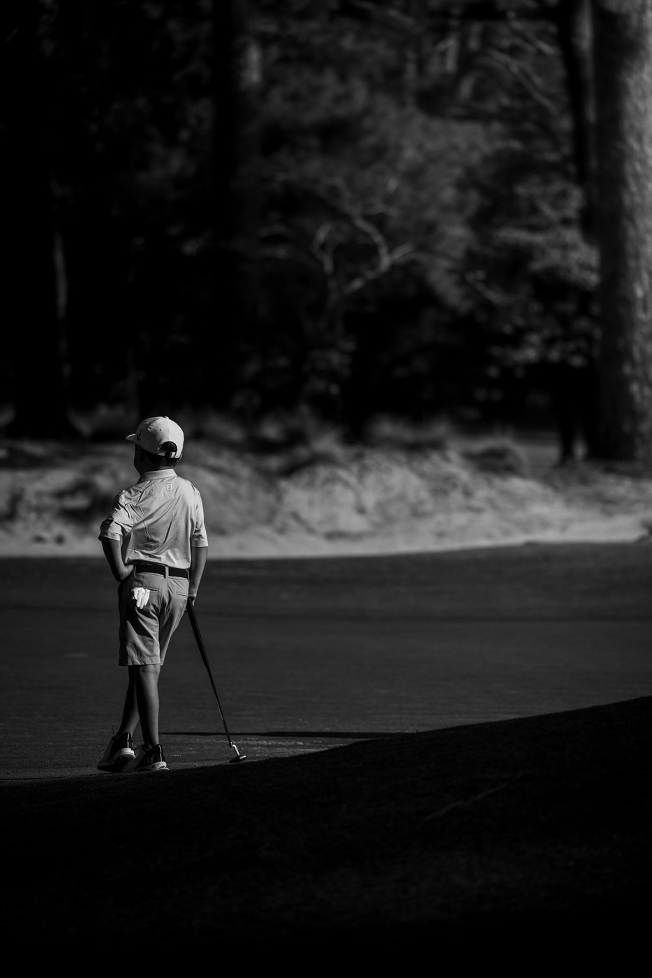 Lone junior golfer standing at the edge of deep shade, black and white