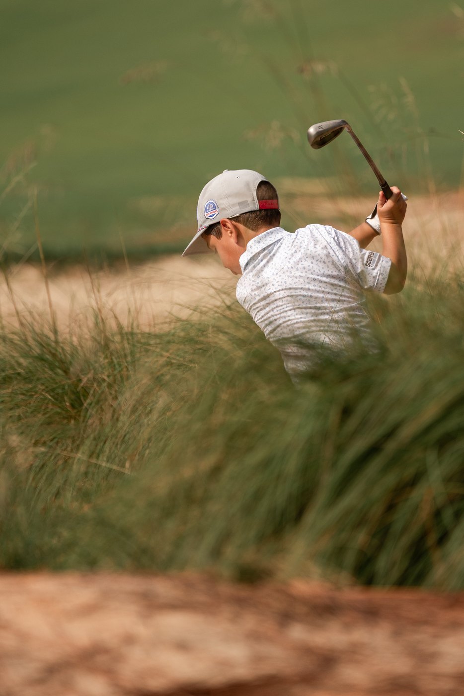 Junior golfer swinging out of a sandy waste area, club at the top, framed by tall wiregrass