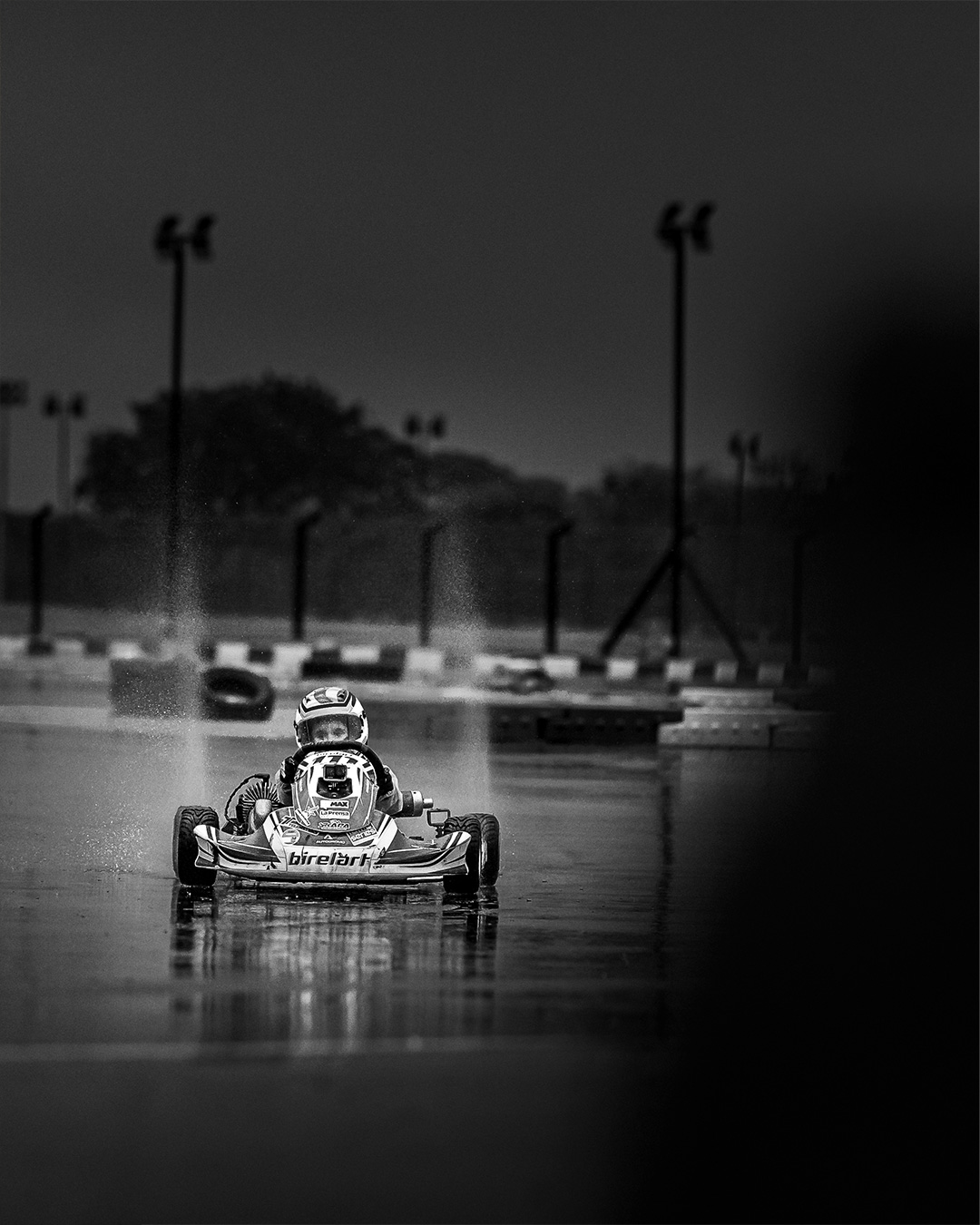 Kart driver head-on through standing water and spray, black and white